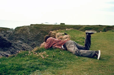 Archibald Robinson lies face down on a grassy coastal cliff wearing a red plaid shirt, grey trousers, and black Converse sneakers.