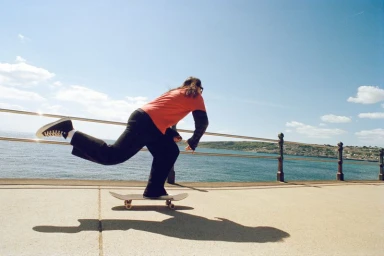 A person skateboarding on a paved seaside promenade overlooking the ocean, wearing a coral red top, dark trousers, and black sneakers.