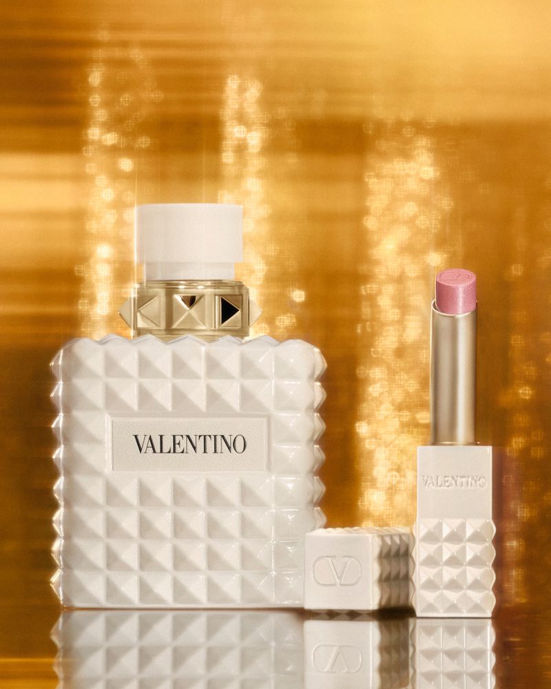 Still life shot of Valentino perfume and lipstick with a textured white design against a golden bokeh background, showcasing beauty products.