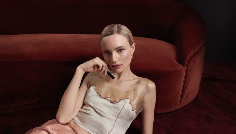 Young woman poses in satin top and pants on the floor next to a velvet couch, holding a tube of lipstick in a beauty shot.