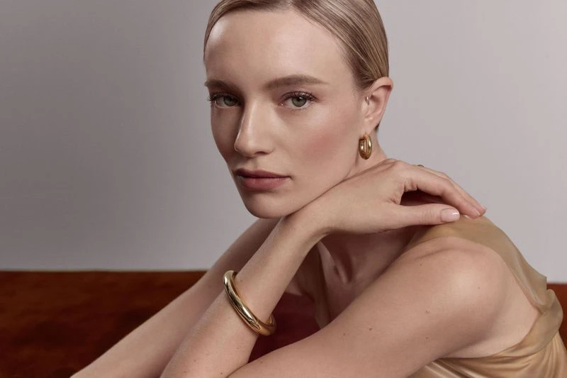 Blonde woman poses indoors wearing gold jewelry, including a hoop earring and a bracelet, in front of a gray and brown background.