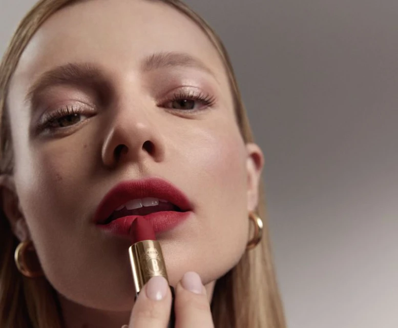 A woman applies red lipstick in a close-up beauty shot against a soft gray background, with gold hoop earrings and light pink makeup.