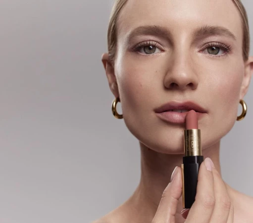 Close-up of a fair-skinned woman applying nude lipstick; she has blonde hair, gold hoop earrings, and a bare shoulder.