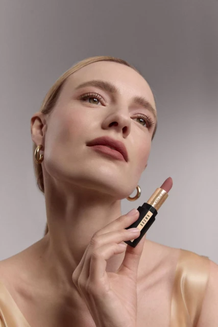 Beauty shot of a woman holding lipstick; neutral makeup, gold hoops, clean light gray background. Beauty and cosmetics product promotion.