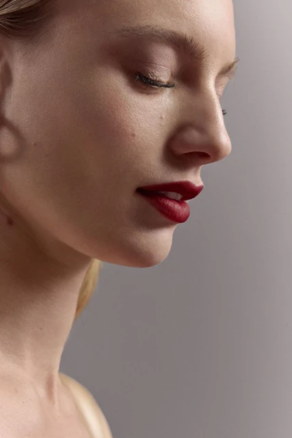 Close-up of a woman's face in profile with red lipstick, fair skin, and closed eyes against a neutral grey background in soft lighting.