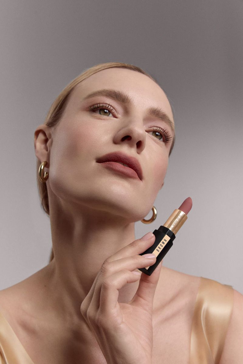 Close-up of a fair-skinned woman with gold hoop earrings holding a lipstick, promoting beauty and cosmetics in a studio setting.