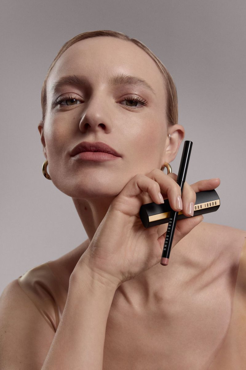 Close-up of a woman with blonde hair and gold hoop earrings, holding Bobbi Brown lip liner and lipstick against a light gray background.