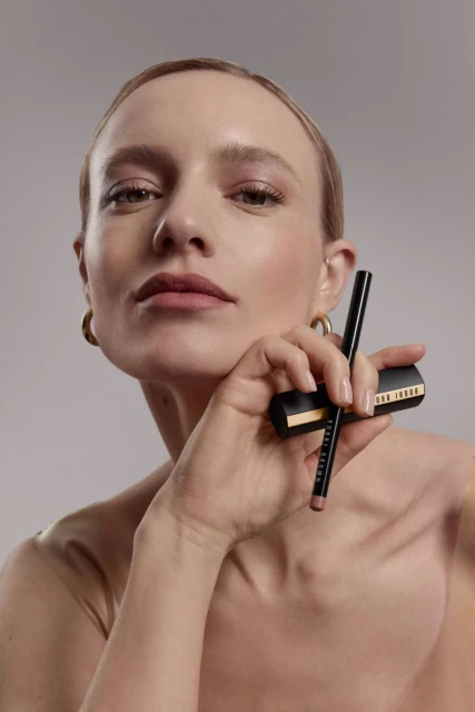 Close-up of a woman with blonde hair and gold hoop earrings, holding Bobbi Brown lip liner and lipstick against a light gray background.