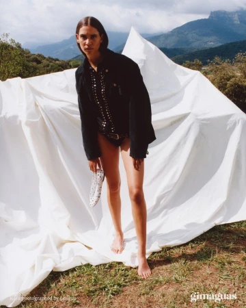 A barefoot woman in jacket and bathing suit poses in front of draped fabric in a grassy field with mountain views on a sunny day.