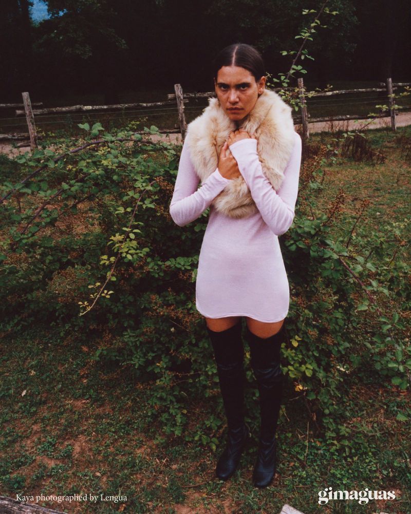 Woman in pink dress and fur stole stands outdoors. Thigh-high boots, dark hair, neutral expression, nature background with wooden fence.