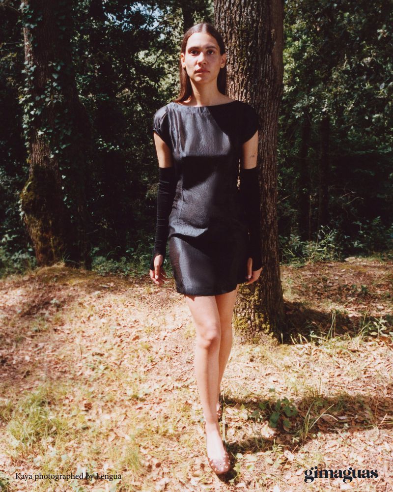 Model Kaya Wilkins wearing a black mini dress and long black arm warmers standing in a sunlit woodland for Gimaguas.