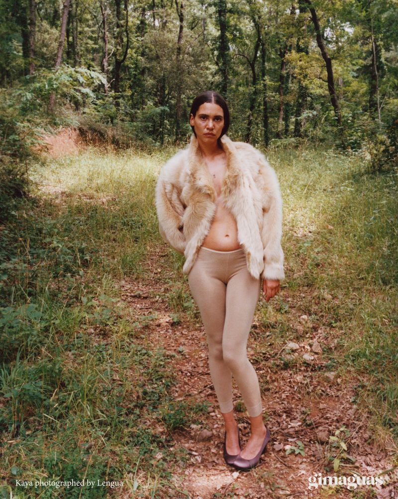 Woman stands in a forest clearing, wearing a light fur coat, leggings, and flats. A dirt path cuts through the green foliage.