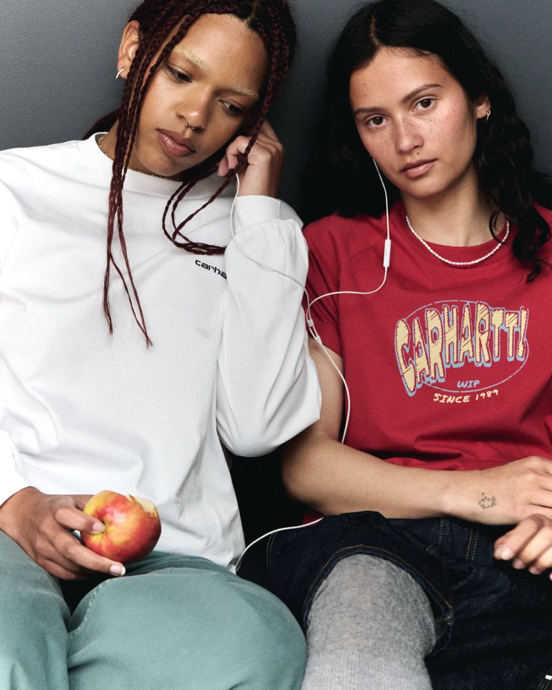 Two young women in casual clothing listen to music with earphones. One holds an apple. The background is a solid gray wall.