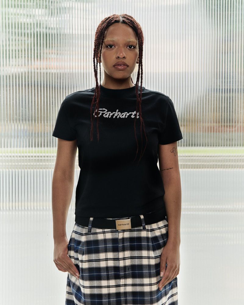 A woman stands indoors wearing a black Carhartt T-shirt, a plaid skirt with a Carhartt belt, and her hair in long braids against a blurred background.