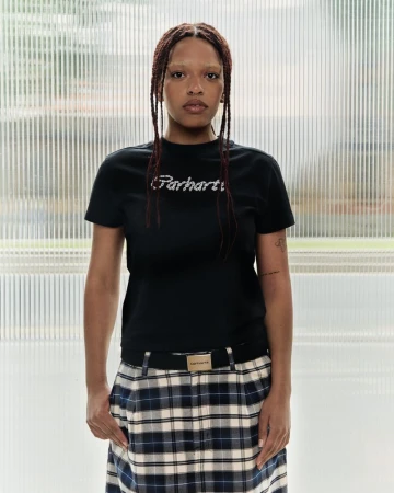 A woman stands indoors wearing a black Carhartt T-shirt, a plaid skirt with a Carhartt belt, and her hair in long braids against a blurred background.