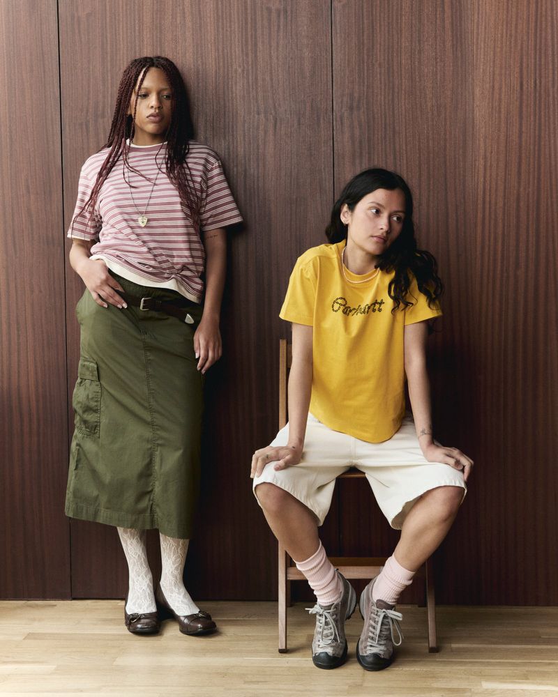Two women in a photo studio wearing casual outfits; one sitting on a wooden chair, the other standing in front of a wood wall.
