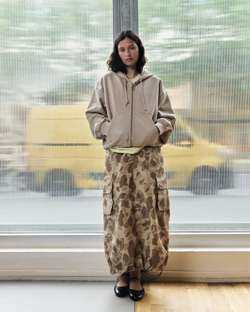 A young woman stands indoors wearing a beige hoodie, patterned maxi skirt, and black flats, in front of a textured window with a yellow vehicle outside.
