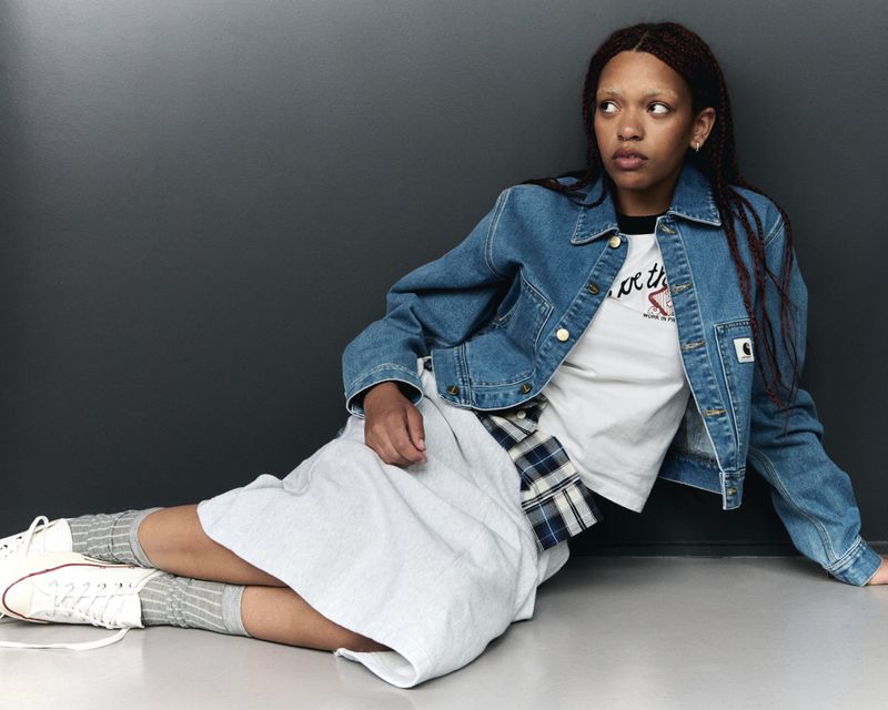 A young adult female sits on the floor in a jean jacket and light-colored skirt, gazing off to the side in a fashion setting.