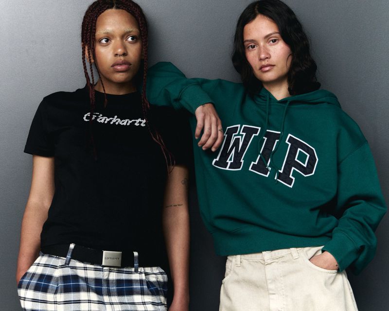 Two young adult women pose indoors against a gray wall, wearing trendy streetwear: a Carhartt t-shirt and WIP hoodie.
