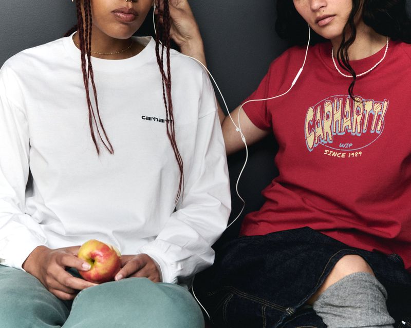 Two young women in casual Carhartt apparel are sitting indoors, connected with earphones, one holding a red apple, with a gray background.