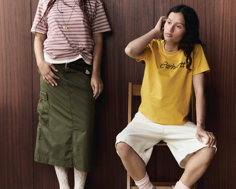 Two women pose in a studio setting with one seated on a chair and wearing a yellow shirt and the other standing with a striped shirt.