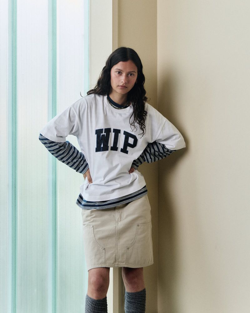 Young woman in white WIP t-shirt and khaki skirt poses indoors with hands on hips in front of a light green partition wall.