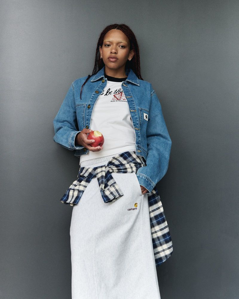 A young adult woman in a denim jacket, white skirt, and plaid shirt tied around her waist holds an apple against a gray background.