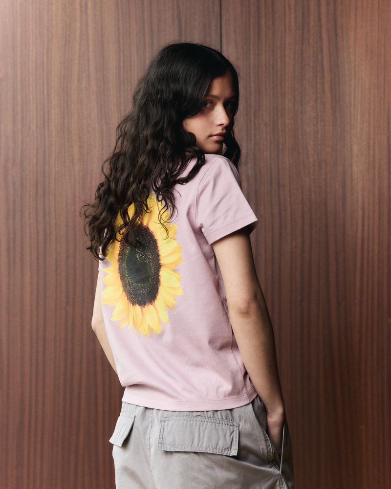 Young woman poses in front of a wood panel wall, wearing a pink t-shirt with a sunflower print and khaki cargo pants.