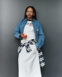 Model wearing a Carhartt denim jacket, white skirt, and plaid shirt tied at the waist holding an apple against a grey backdrop.