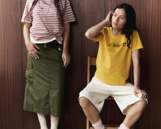Two models posing against a dark wood-paneled wall wearing Carhartt S/S 25 streetwear apparel including a yellow t-shirt and cargo skirt.