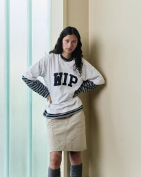 Model wearing white WIP graphic t-shirt and beige utility skirt standing indoors against a pale wall.