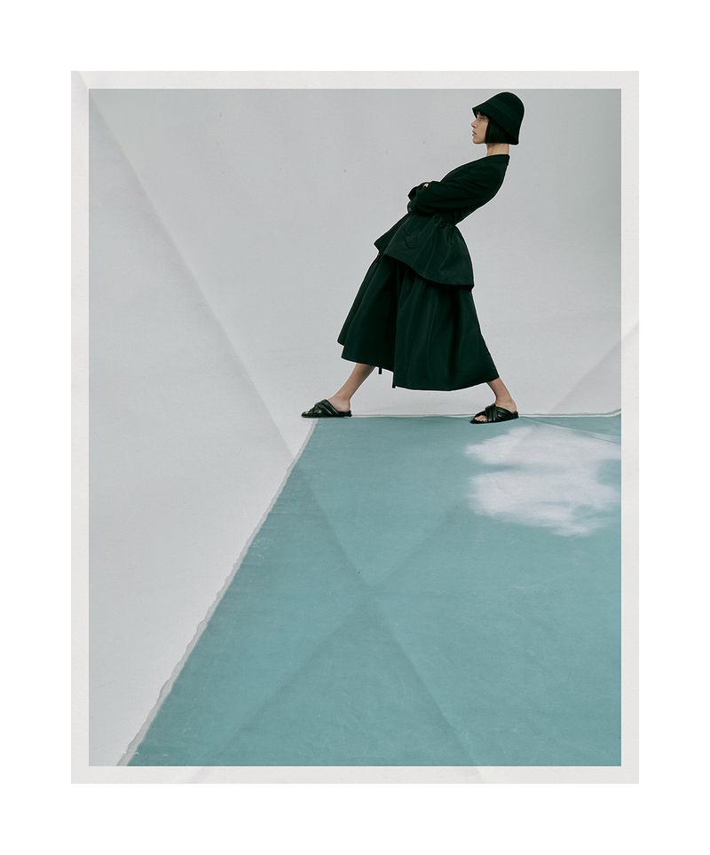 Fashion shot of an adult female model in a black dress and bucket hat, posing on a blue geometric platform against a white backdrop.