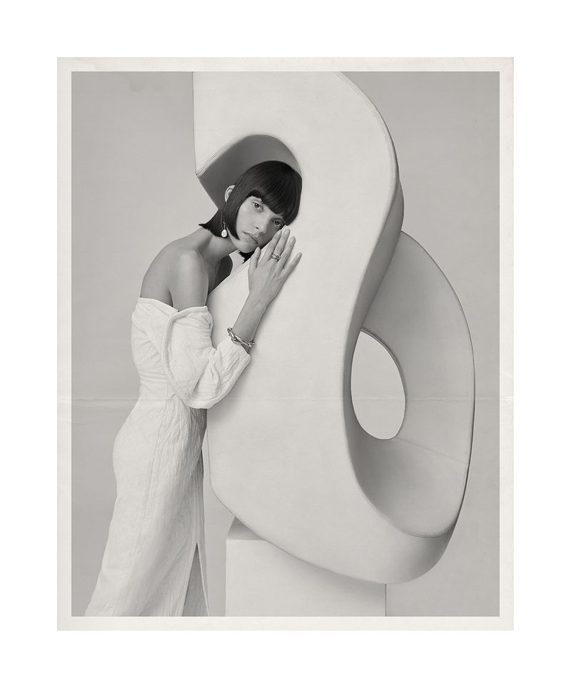 Monochrome portrait of a woman with a blunt bob, leaning on a white abstract sculpture in a textured off-the-shoulder dress with a slit.