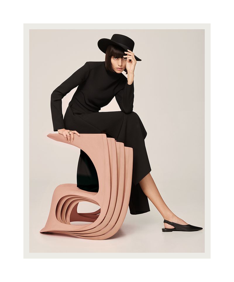 Woman in black outfit and hat seated on pink modern art chair in a studio setting, showcasing fashion and creative design.