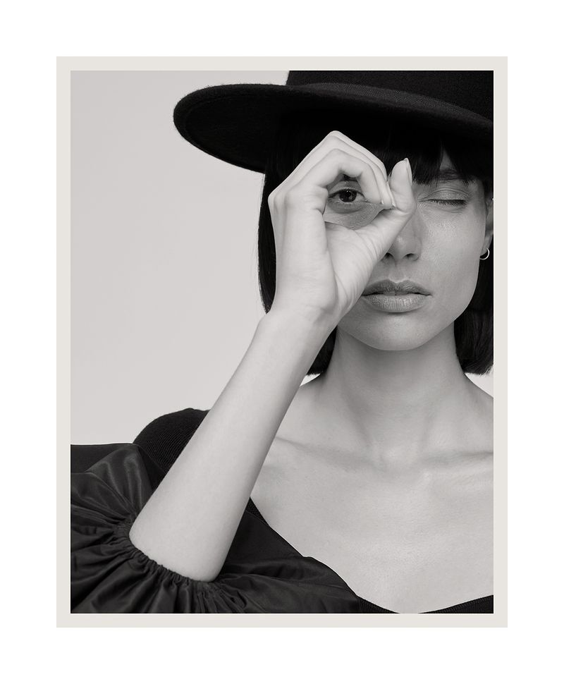 Monochrome portrait of a woman wearing a black hat, glancing at the camera, making an 'OK' sign with her hand over one eye.