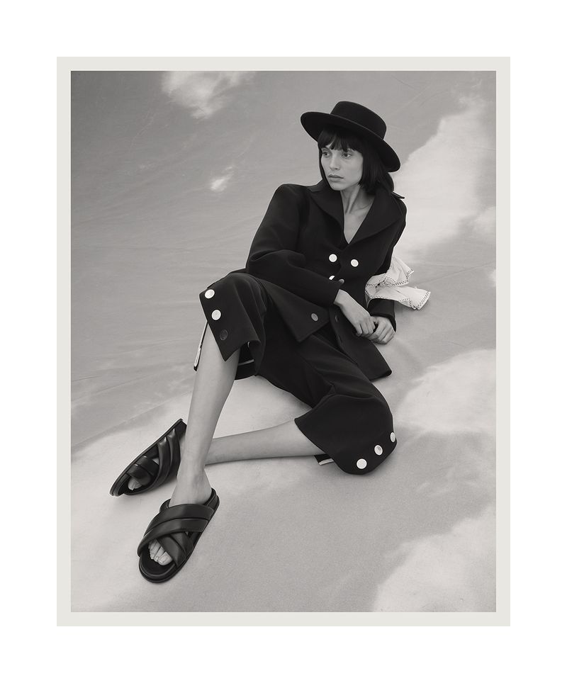 Monochrome fashion shot of a woman in a dark suit and hat, seated in a cloud-like setting, showcasing modern editorial style.