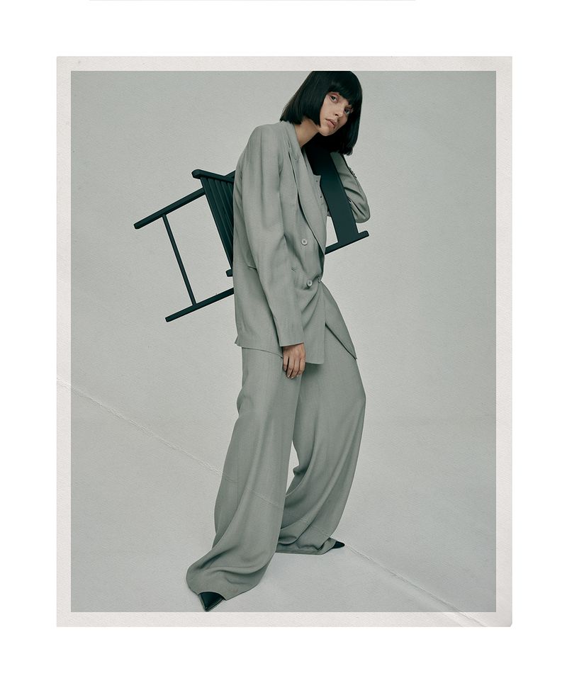 Adult woman in a grey suit posing in front of a grey background with a black chair and black pointed-toe heels, studio setting.