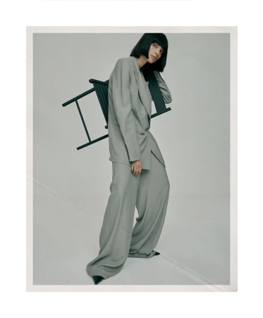 Adult woman in a grey suit posing in front of a grey background with a black chair and black pointed-toe heels, studio setting.