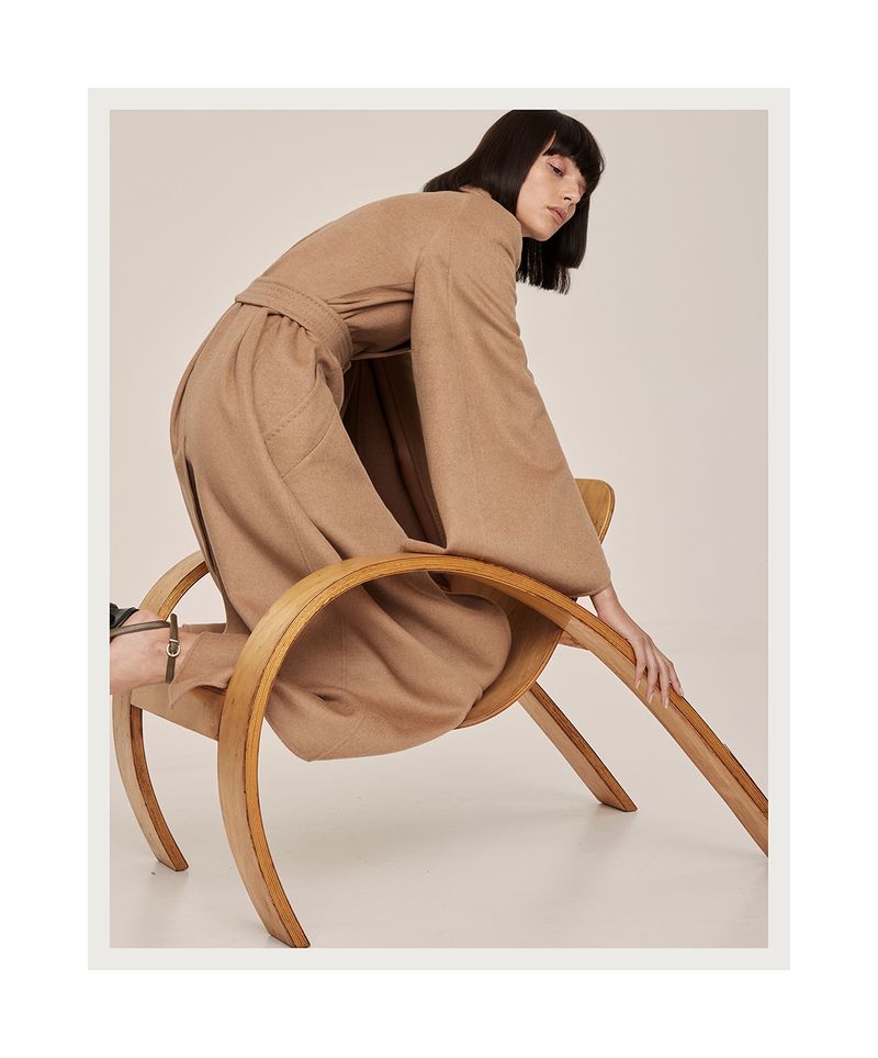 Fashion shot of a brunette woman in a tan outfit kneeling on a wooden chair. The image is well-lit and creates a stylish, modern aesthetic.