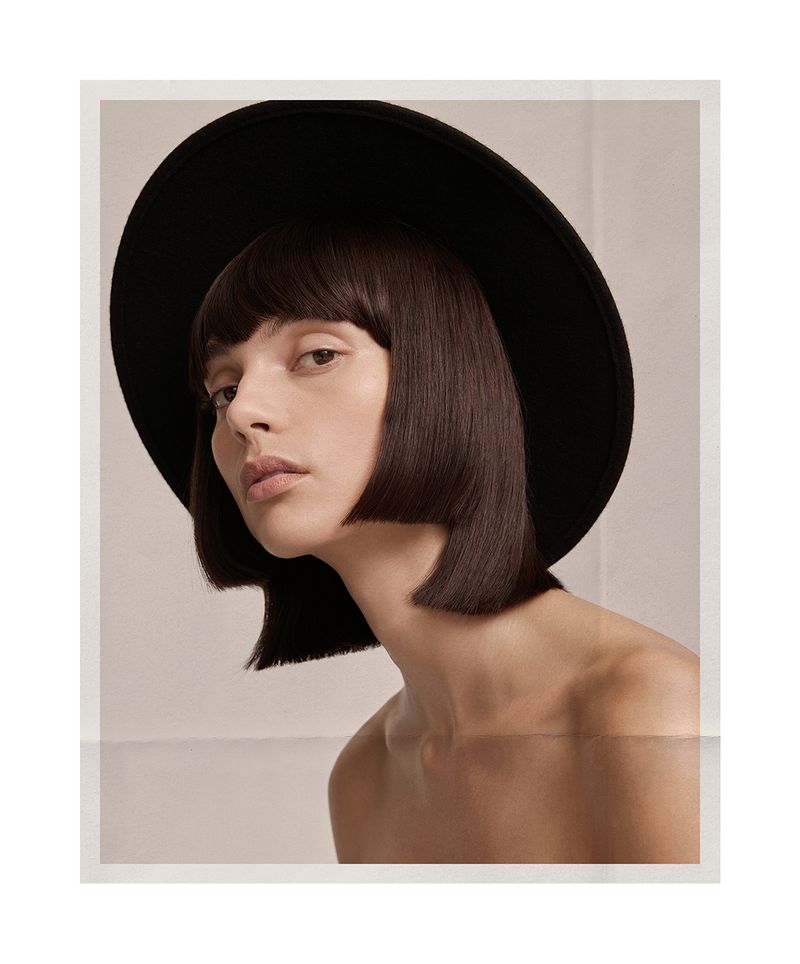 Close-up portrait of model Charlee Fraser wearing a black wide-brim hat and blunt bob haircut against a white studio background.