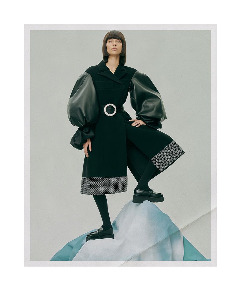 Fashionable woman in a dark coat with oversized sleeves posing on a geometric backdrop. The studio shot highlights the coat's modern design.