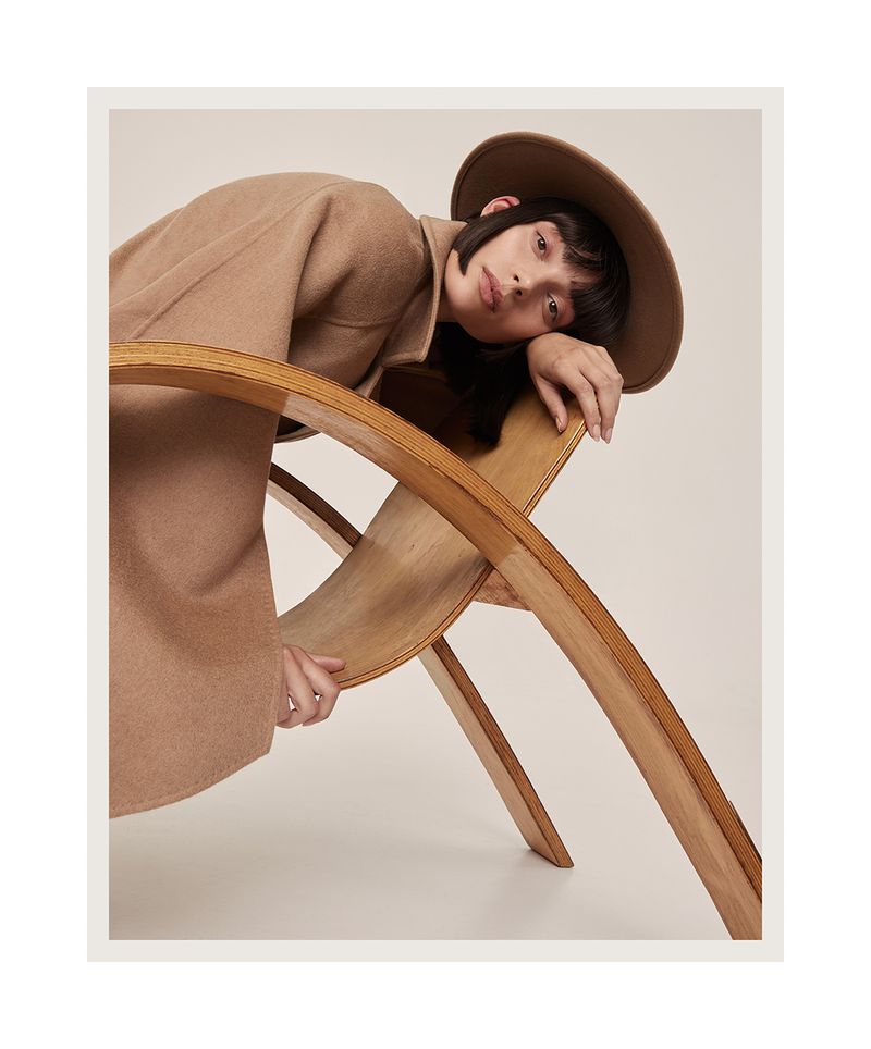 Stylish woman in a tan coat and hat poses in a photo studio, leaning against a bentwood chair, in a fashion editorial style.