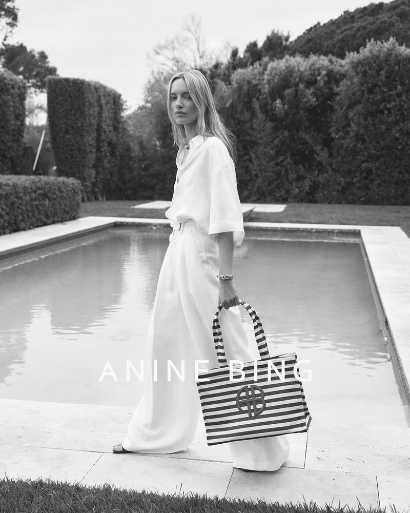 Elegant blonde woman in white linen holding striped tote bag next to pool, wearing wide leg pants and long sleeve shirt, outdoor setting.