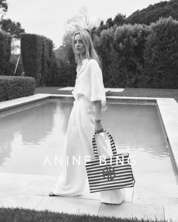 Elegant blonde woman in white linen holding striped tote bag next to pool, wearing wide leg pants and long sleeve shirt, outdoor setting.
