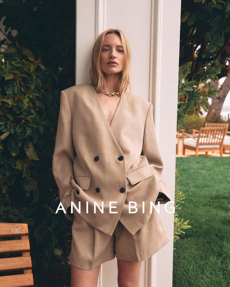 A stylish woman in a beige blazer and shorts stands outdoors near greenery, showcasing a modern and sophisticated fashion aesthetic.
