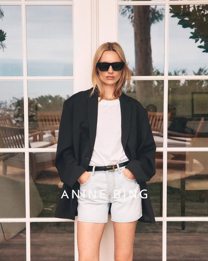 Blonde woman in sunglasses, white tee, light wash denim shorts, and black blazer poses in front of white French doors.
