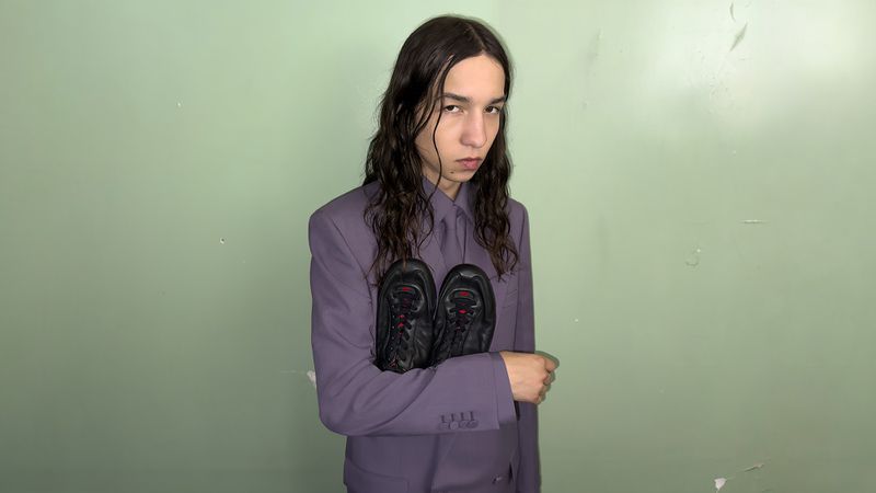 Person in purple suit holding pair of black sneakers with red laces in indoor shot against a green wall. Fashion portrait.