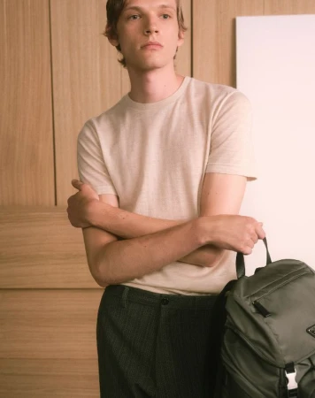 A young adult male stands indoors with arms crossed, wearing a t-shirt and trousers, holding a Prada backpack against a wood-panelled wall.