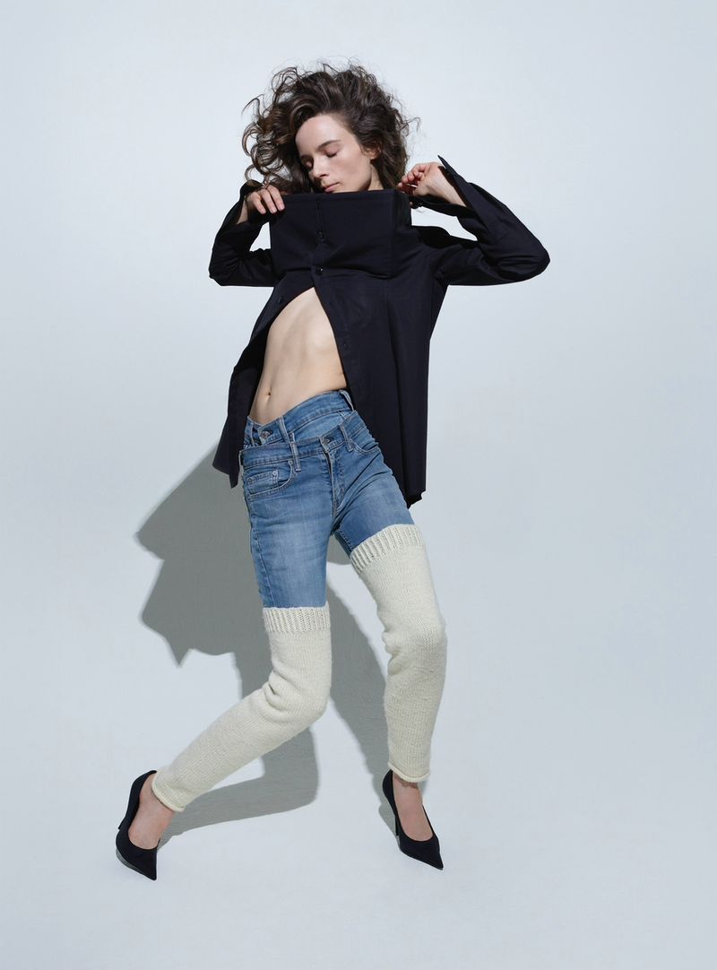 Fashion shot of a woman in denim jeans and a black jacket, posing against a white backdrop in black high heels with leg warmers.