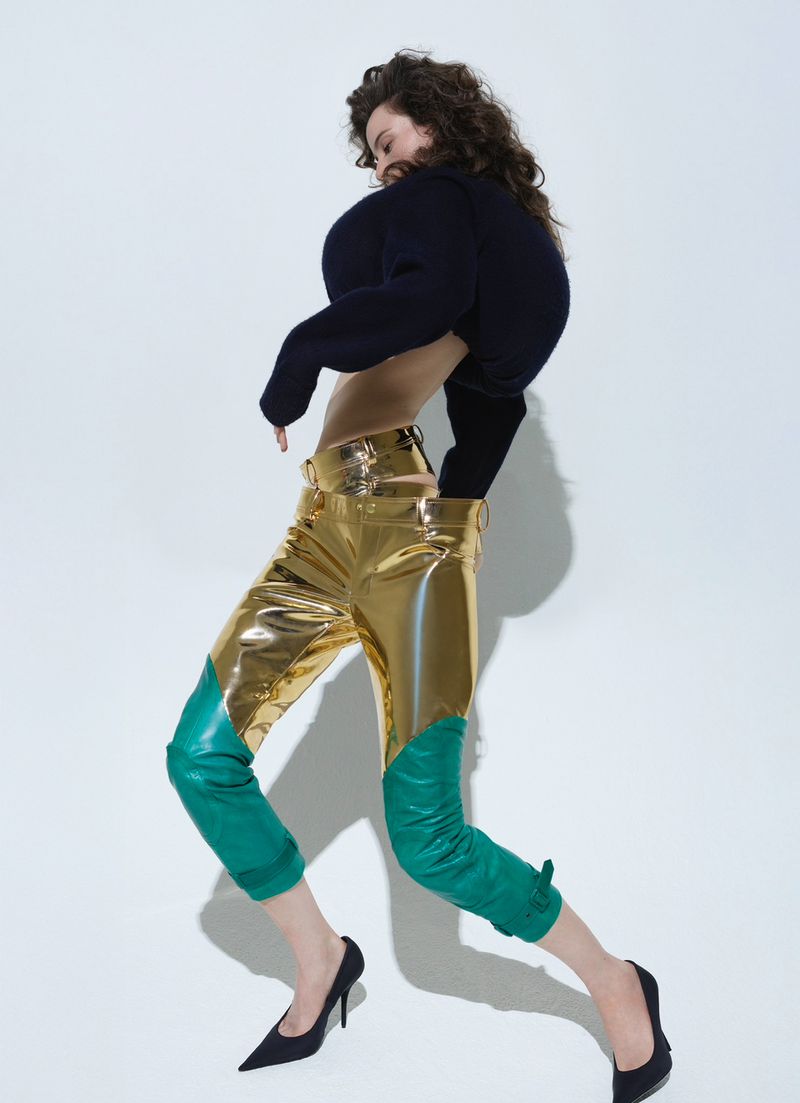 A woman with curly hair poses in a studio wearing a navy sweater and bright gold and teal-colored leather trousers and black high heels.